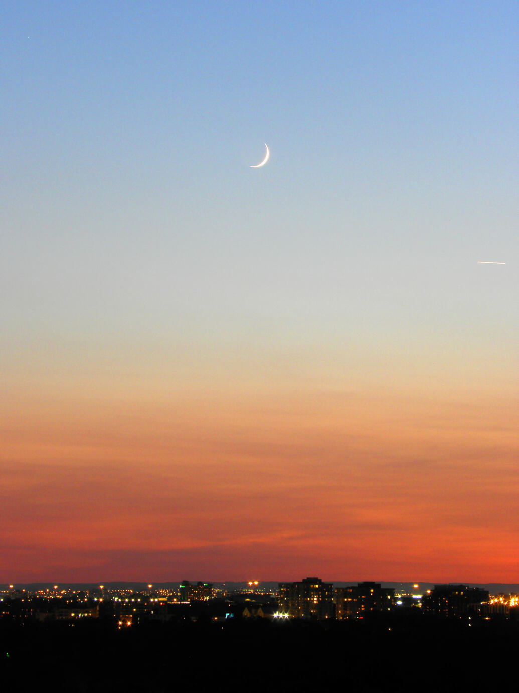 A slender crescent Moon floats above Toronto as twilight settles over the city. Warm city lights shimmer beneath a sky fading from soft blue to deep amber, creating a quiet and dreamlike evening scene.