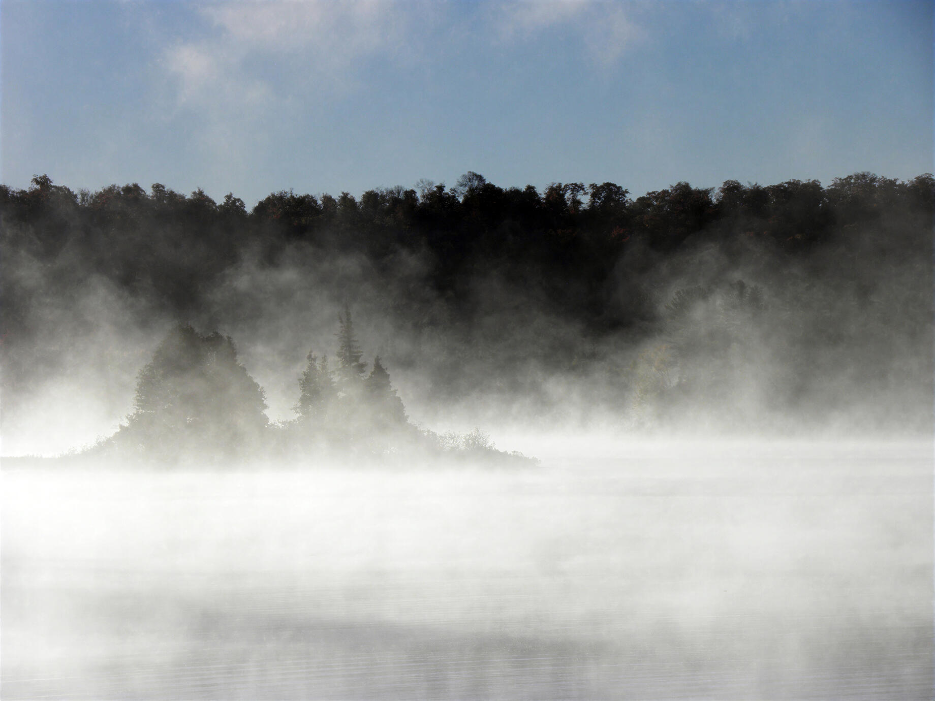 Morning mist drifts across Dwight Bay, veiling the shoreline and trees in a soft, ghostly haze. The still water and muted autumn landscape create a quiet, dreamlike atmosphere.